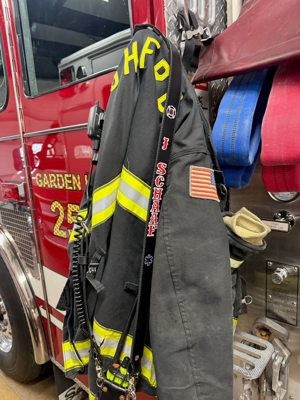 A Box 31 hand-painted radio strap is shown hanging with turnout gear on the door of a fire engine in the Garden Homes Fire Protection District in the Chicagoland area. This in-service strap is black leather with red letters and white outlines and has Box 31's universal radio bucket.