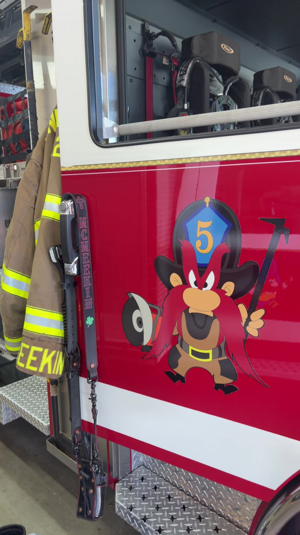 A Box 31 hi-def radio strap, with black text and red outlines printed directly to leather, hangs on the door of a fire engine in western Pennsvylvania. The engine has a large Yosemite Sam on the door, holding a halligan and a K12 and with a blue shield bearing the number 5. The radio strap has a cross at the top, followed by MCMEEKIN, with a green four-leaf clover below the name.