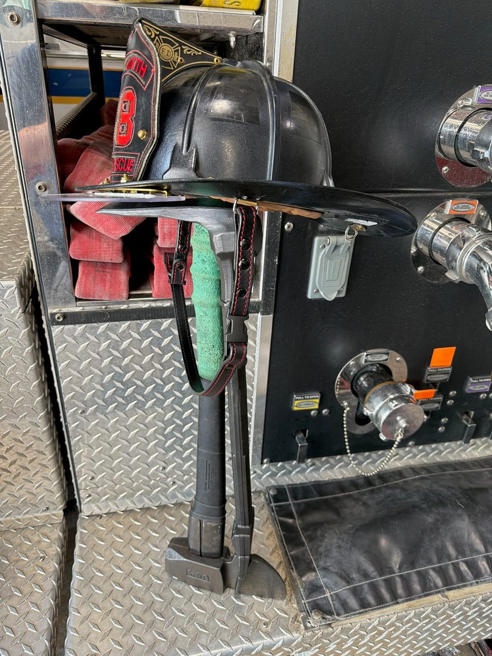 A black leather Box 31 quick release chin strap, with red stitching, is in service and staged on top of a set of irons by the hose bed on a fire engine.