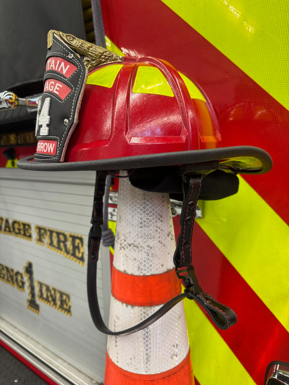 A Box 31 black leather chin strap with red stitching hangs on a red fire helmet, which is sitting atop a traffic cone beside Engine 1 at a station in Minnesota.
