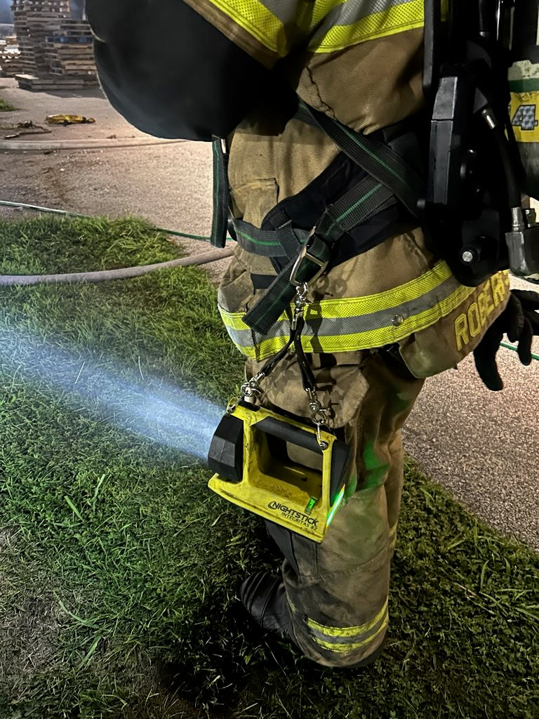 A firefighter in full turnout gear has a box light flashlight clipped to his gear with a Box 31 Box Light Clip, and a beam of light shines across the yard.