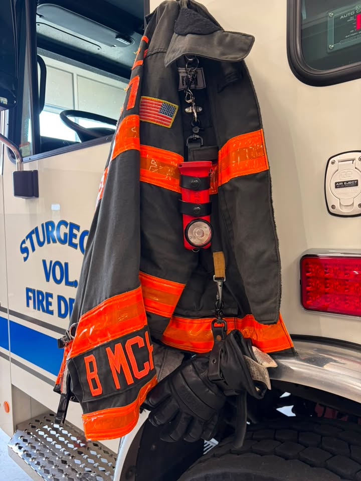 Black leather inverted flashlight holder from Box 31 is clipped to a black bunker coat with orange reflective trim, hanging on the door of a white fire engine from Sturgeon Volunteer Fire Department.