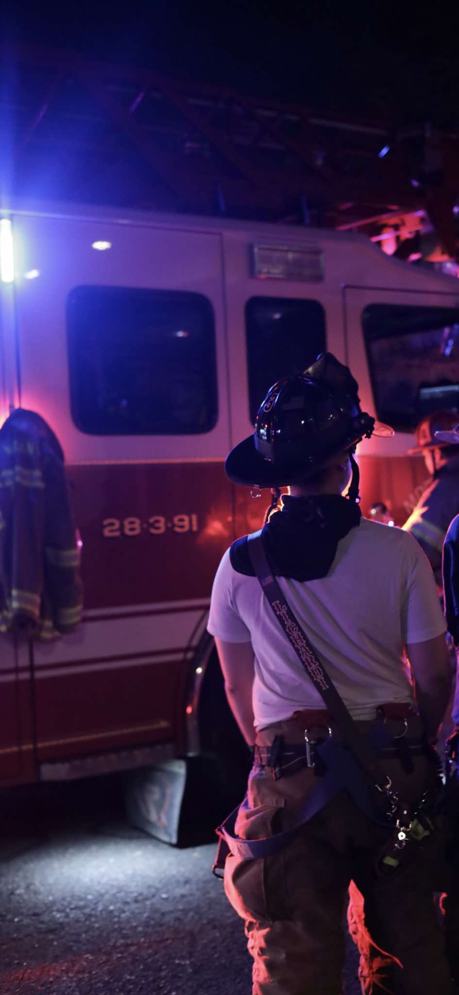 A firefighter in New Jersey wears a hand-painted Box 31 radio strap, standing beside a fire engine at night, with the scene lit by emergency lights.