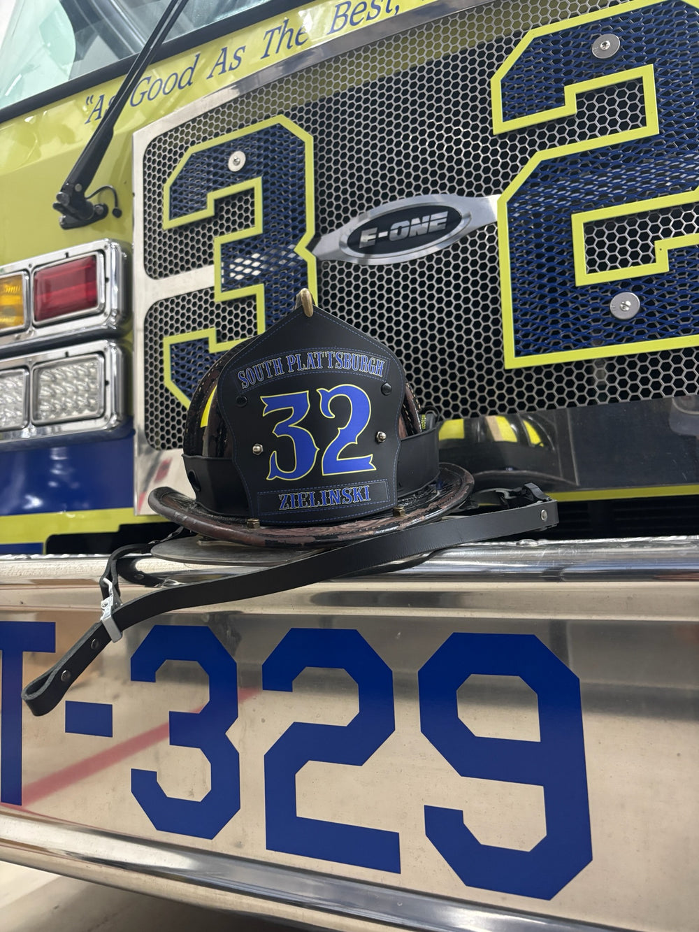 This Box 31 traditional-style shield is in service in upstate New York. Shield is black leather with blue text and yellow outlines, staged on the front bumper of a yellow fire truck.