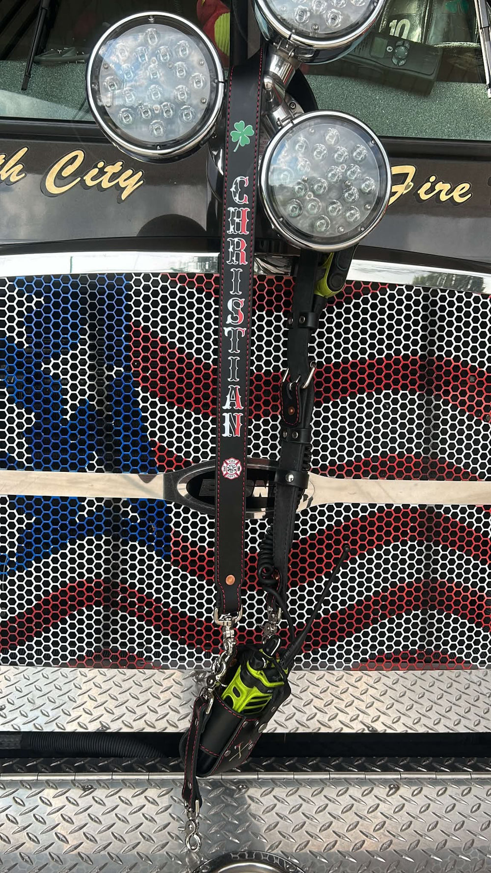 Box 31 hi-def black leather radio strap hanging from the spinner on the front of a fire engine with an American flag grille. The strap has CHRISTIAN printed vertically in Box 31’s Thin Red Line design, with white lettering accented in red.