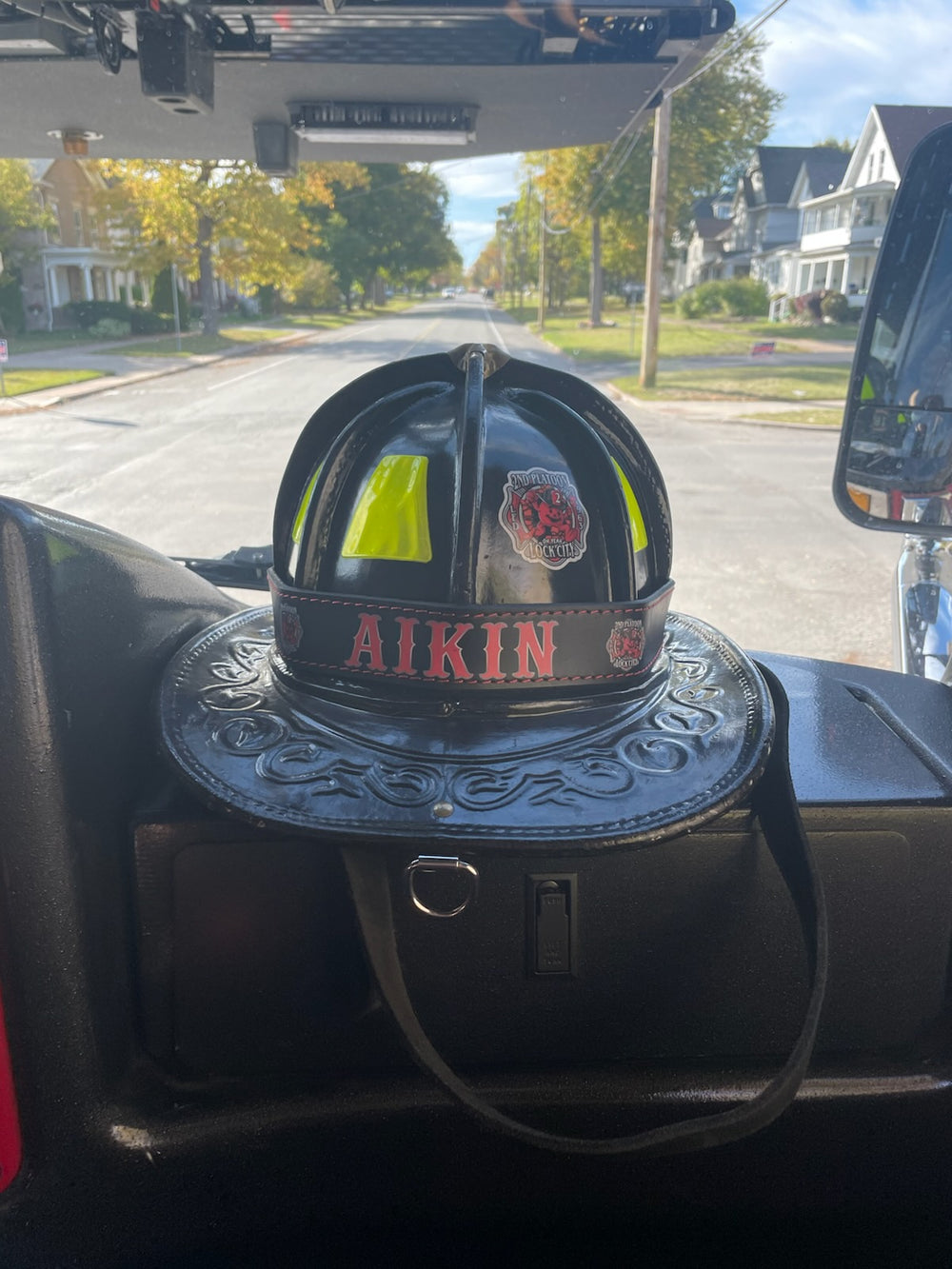 A hi-def printed helmet band, black leather with red lettering, is shown on a fire helmet, on the dash of a truck driving down a residential street in New York State.